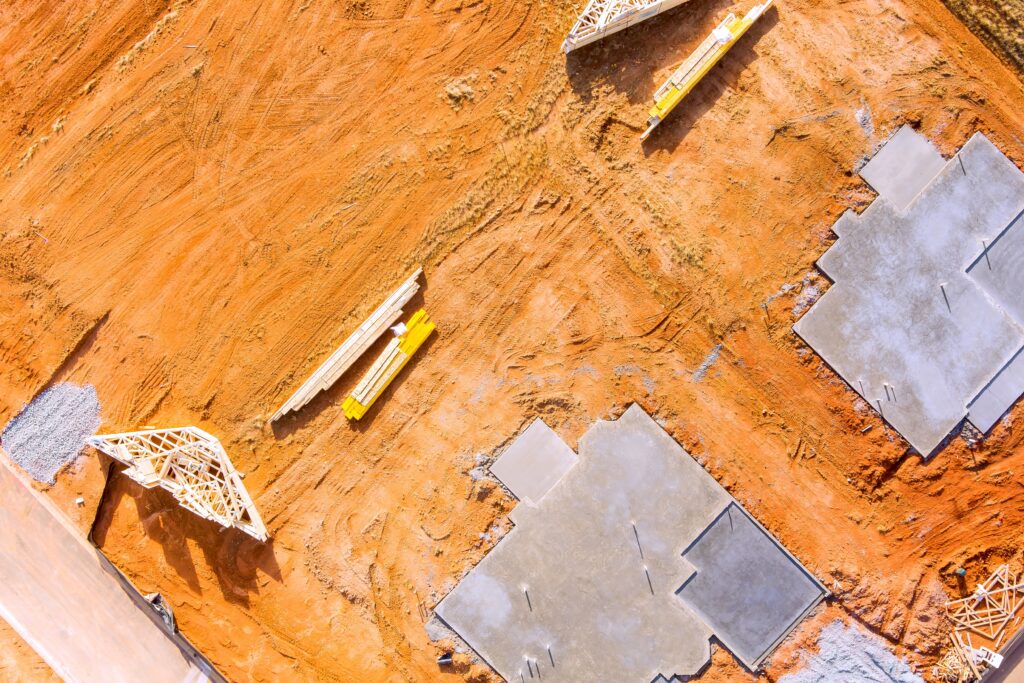 An aerial view of a home lot construction site featuring two concrete building foundations and scattered wooden framing materials on red dirt.